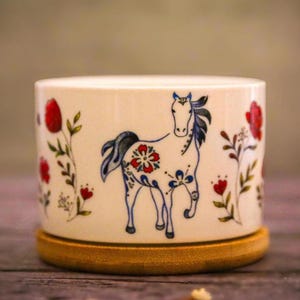 May include: A white ceramic plant pot featuring a hand-painted horse design. The horse is decorated with red and blue floral patterns. Red flowers and green leaves are painted around the pot. It sits on a round, wooden coaster.