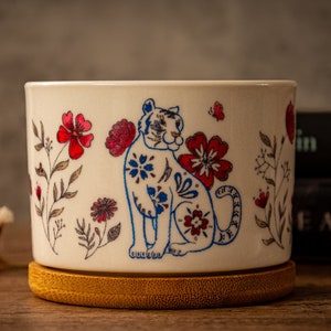 May include: White ceramic planter with a blue and white tiger design and red floral accents. The planter has a bamboo base.