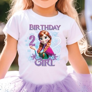 May include: A young girl wearing a white t-shirt with a graphic of a cartoon character and the words "BIRTHDAY 2 GIRL". She is also wearing a purple tutu and purple sunglasses. The background is blurred, suggesting an outdoor party setting.