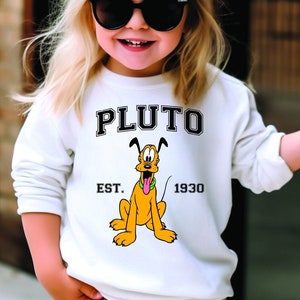 Puede incluir: Una sudadera blanca con una imagen de dibujos animados de Pluto el perro. La sudadera tiene el texto "PLUTO" en letras negras sobre la imagen y "EST. 1930" en letras negras debajo de la imagen.