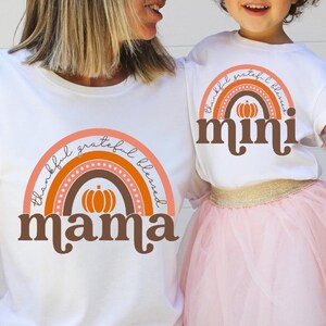 May include: White t-shirts with matching rainbow designs. The adult shirt says "mama" and the child's shirt says "mini". Both shirts have a pumpkin graphic and the words "thankful, grateful, blessed". The child is wearing a pink tutu.