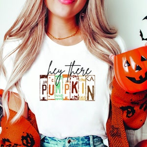 May include: White t-shirt with the text "hey there PUMPKIN" in a decorative font. The word "PUMPKIN" is spelled out using license plates. An orange pumpkin-shaped mug with a green handle is held in the hand.
