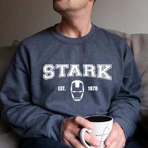 Pode incluir: Um moletom azul marinho com a palavra "STARK" em letras brancas, juntamente com "EST. 1970" e um gr&aacute;fico do capacete do Homem de Ferro. A pessoa est&aacute; segurando uma caneca branca com interior preto e design geom&eacute;trico.