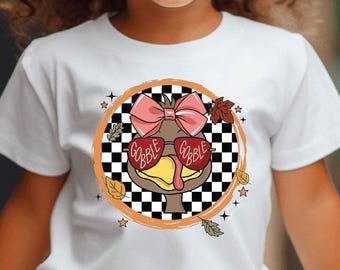 May include: A white t-shirt featuring a cartoon turkey wearing sunglasses with the words "Gobble Gobble" and a pink bow. The design is set within a black and white checkered circle, with fall leaves and stars around it.