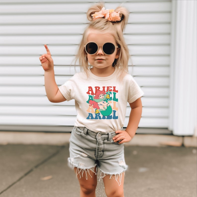 May include: A young girl wearing a white t-shirt with a colourful graphic of Ariel the Little Mermaid and the text "Ariel" printed on it. She is also wearing denim shorts and sunglasses.