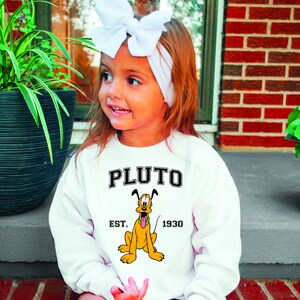 Puede incluir: Sudadera blanca con una imagen de dibujos animados de Pluto, el perro de Disney. La sudadera tiene el texto "PLUTO EST. 1930" impreso en ella.