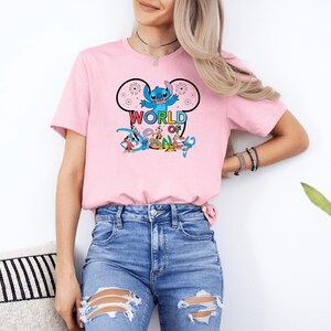 Pu&ograve; includere: Una maglietta rosa chiaro con una grafica a cartoni animati di Stitch di Lilo & Stitch, con le parole "WORLD OF" sopra e "DREAM" sotto. Il design include altri personaggi dei cartoni animati. La maglietta &egrave; abbinata a jeans blu.