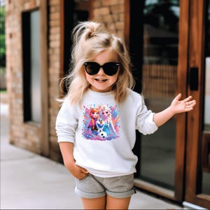 May include: A white long-sleeved sweatshirt featuring a colorful graphic of characters from the movie Frozen. The graphic includes Anna, Elsa, and Olaf. The sweatshirt is worn by a young child wearing sunglasses and gray shorts.