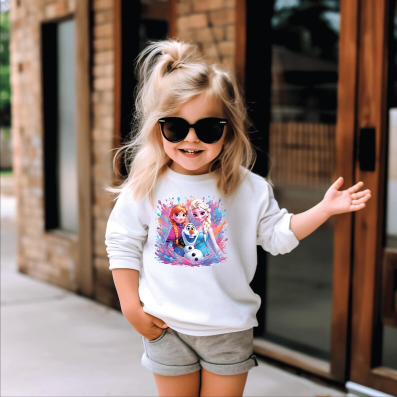 May include: A white long-sleeved sweatshirt featuring a colorful graphic of characters from the movie Frozen. The graphic includes Anna, Elsa, and Olaf. The sweatshirt is worn by a young child wearing sunglasses and gray shorts.