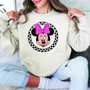 Puede incluir: Una sudadera blanca con un dise&ntilde;o circular de cuadros negros y blancos que presenta una imagen de dibujos animados de Minnie Mouse con un lazo rosa.