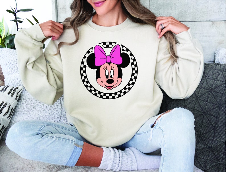 Puede incluir: Una sudadera blanca con un dise&ntilde;o circular de cuadros negros y blancos que presenta una imagen de dibujos animados de Minnie Mouse con un lazo rosa.