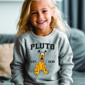 Puede incluir: Una sudadera gris con una imagen de dibujos animados de Pluto, el perro de Disney. La sudadera tiene el texto "PLUTO" en letras negras sobre la imagen y "EST. 1930" en letras negras debajo de la imagen.