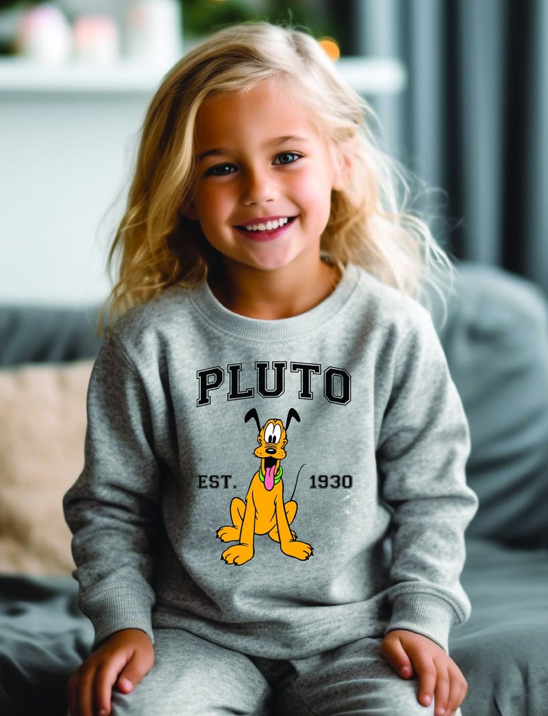 Puede incluir: Una sudadera gris con una imagen de dibujos animados de Pluto, el perro de Disney. La sudadera tiene el texto "PLUTO" en letras negras sobre la imagen y "EST. 1930" en letras negras debajo de la imagen.