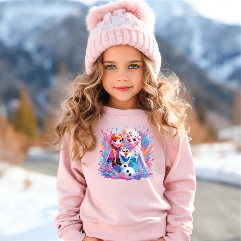 May include: A young girl wearing a light pink sweatshirt and a matching pink knit hat with a pom-pom. The sweatshirt features a colorful graphic of Anna, Elsa, and Olaf from the movie Frozen. The background shows a snowy mountain scene.
