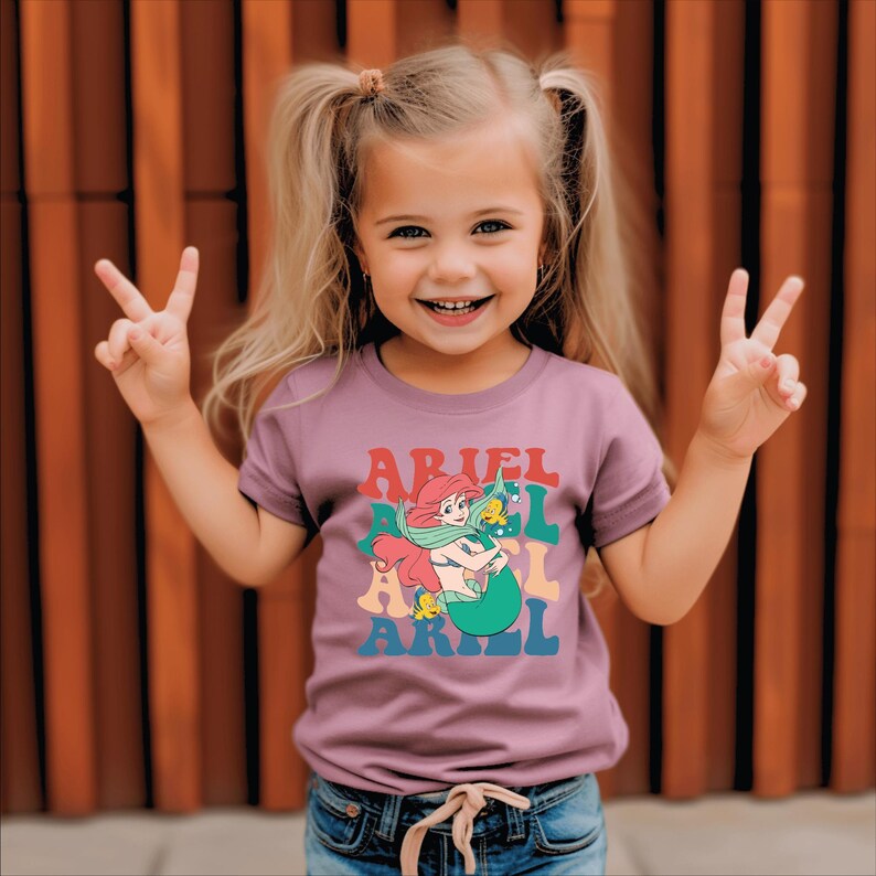 May include: A young girl wearing a purple t-shirt with a retro-style graphic of Ariel the Little Mermaid. The graphic features Ariel in a rainbow-coloured font with the text "Ariel" repeated multiple times.