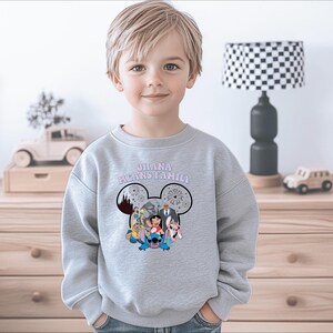 K&ouml;nnte beinhalten: Ein hellgraues Sweatshirt mit einem Mickey Mouse-Kopf-Design und den Worten "OHANA MEANS FAMILY" in Rosa. Das Design beinhaltet Zeichentrickfiguren. Das Sweatshirt wird von einem kleinen Kind getragen.