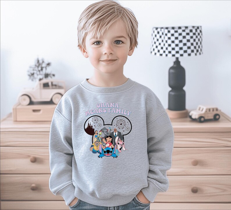 K&ouml;nnte beinhalten: Ein hellgraues Sweatshirt mit einem Mickey Mouse-Kopf-Design und den Worten "OHANA MEANS FAMILY" in Rosa. Das Design beinhaltet Zeichentrickfiguren. Das Sweatshirt wird von einem kleinen Kind getragen.