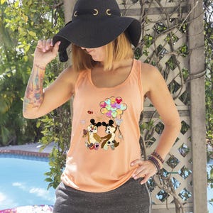 May include: Peach-colored tank top featuring a cartoon graphic of Chip and Dale holding hands with balloons. The tank top is paired with gray shorts and a black sun hat with gold accents. The person is wearing multiple bracelets.