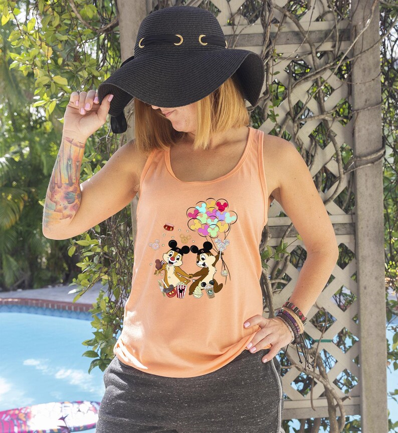 May include: Peach-colored tank top featuring a cartoon graphic of Chip and Dale holding hands with balloons. The tank top is paired with gray shorts and a black sun hat with gold accents. The person is wearing multiple bracelets.