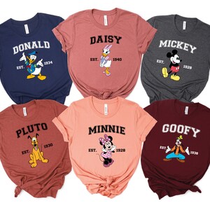 May include: Six different colored t-shirts with Disney character graphics and text. The shirts are all in a similar style with a crew neck and short sleeves. The text on the shirts reads "Donald", "Daisy", "Mickey", "Pluto", "Minnie", and "Goofy".
