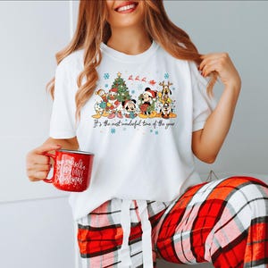 May include: White t-shirt featuring a colorful graphic of Disney characters, a Christmas tree, and the text "It's the most wonderful time of the year." The person is holding a red mug with white text. The person is wearing red and white plaid pajama pants.