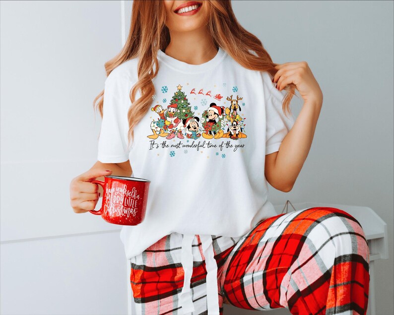 May include: White t-shirt featuring a colorful graphic of Disney characters, a Christmas tree, and the text "It's the most wonderful time of the year." The person is holding a red mug with white text. The person is wearing red and white plaid pajama pants.