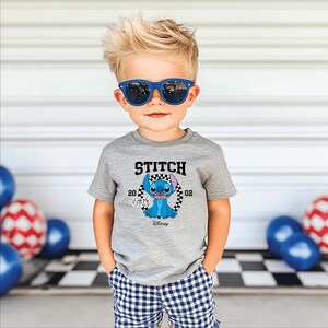 May include: A young child wearing blue sunglasses and a gray t-shirt with a Stitch graphic. The shirt features the word "STITCH" above a cartoon image of Stitch, with the numbers "20" and "02" on either side. The child is also wearing blue and white checkered shorts.