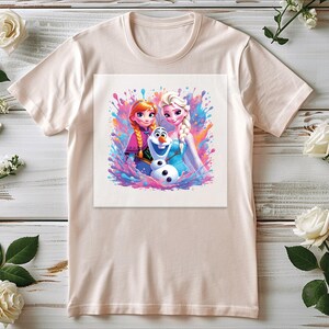 Pu&ograve; includere: Una maglietta beige chiaro con una grafica colorata dei personaggi animati Anna, Elsa e Olaf del film Frozen. La grafica ha un effetto acquerello con spruzzi di rosa, blu e viola.