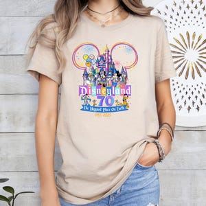 May include: Beige t-shirt featuring a colorful graphic celebrating Disneyland's 70th anniversary. The design includes the Disneyland castle, Mickey Mouse ears, and various Disney characters. Text reads "Disneyland 70 The Happiest Place On Earth 1955-2025".
