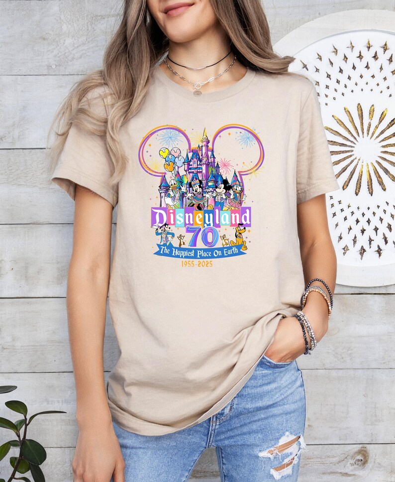 May include: Beige t-shirt featuring a colorful graphic celebrating Disneyland's 70th anniversary. The design includes the Disneyland castle, Mickey Mouse ears, and various Disney characters. Text reads "Disneyland 70 The Happiest Place On Earth 1955-2025".