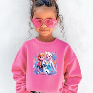 May include: A young girl wearing a pink sweatshirt with a graphic of Anna, Elsa, and Olaf from Frozen. She is also wearing pink heart-shaped sunglasses and blue jeans. Her hair is styled in a bun.