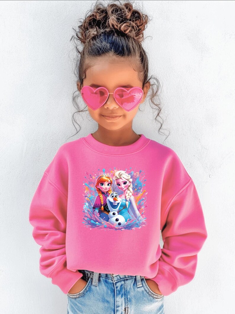 May include: A young girl wearing a pink sweatshirt with a graphic of Anna, Elsa, and Olaf from Frozen. She is also wearing pink heart-shaped sunglasses and blue jeans. Her hair is styled in a bun.