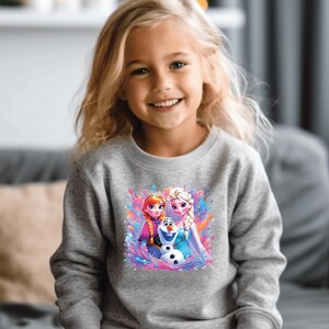 May include: A young girl wearing a gray sweatshirt with a colorful graphic of animated characters. The graphic features two princesses and a snowman against a vibrant splash background. The girl is smiling.