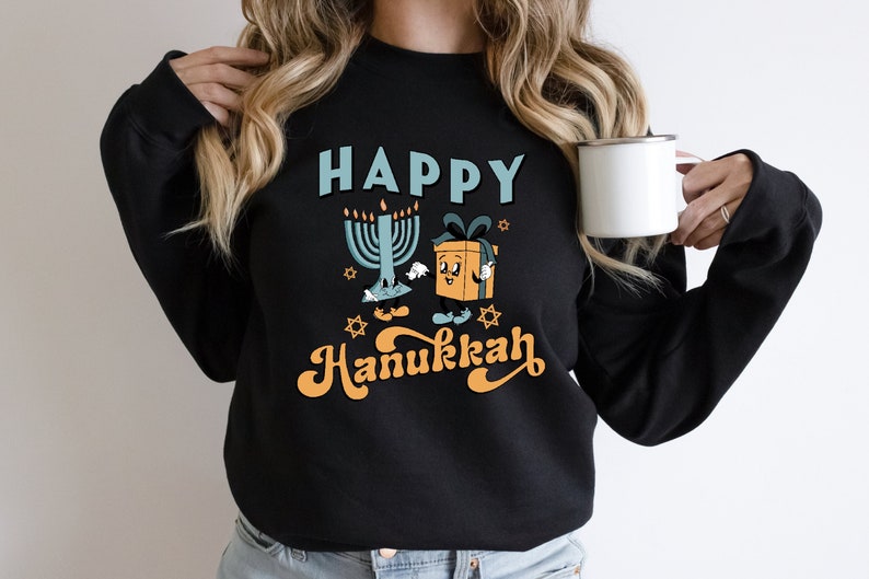 May include: Black sweatshirt with a colorful graphic design. The design features the words "HAPPY Hanukkah" in blue and orange, along with illustrations of a menorah and a gift box. The sweatshirt is a comfortable, casual style.
