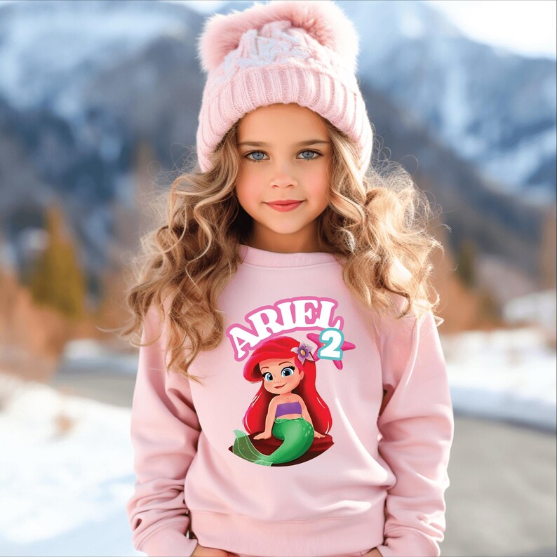 Może przedstawiać: Młoda dziewczyna w jasnor&oacute;żowej bluzie z grafiką Małej Syrenki Arielki i cyfrą 2. Ma r&oacute;wnież na sobie jasnor&oacute;żową dzianinową czapkę z pomponem. Tło to rozmyty widok na zaśnieżoną g&oacute;rę.