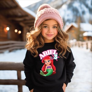 Może przedstawiać: Młoda dziewczyna w czarnej bluzie z grafiką Arielki z Małej Syrenki i cyfrą 3. Ma r&oacute;wnież na sobie r&oacute;żową dzianinową czapkę. Tło przedstawia zaśnieżony krajobraz i drewniane budynki.