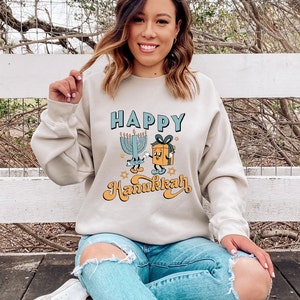 May include: Beige sweatshirt with the words "HAPPY Hanukkah" in blue and orange lettering. The graphic includes a cartoon menorah, a gift box, and stars. The person is wearing blue jeans and white sneakers.