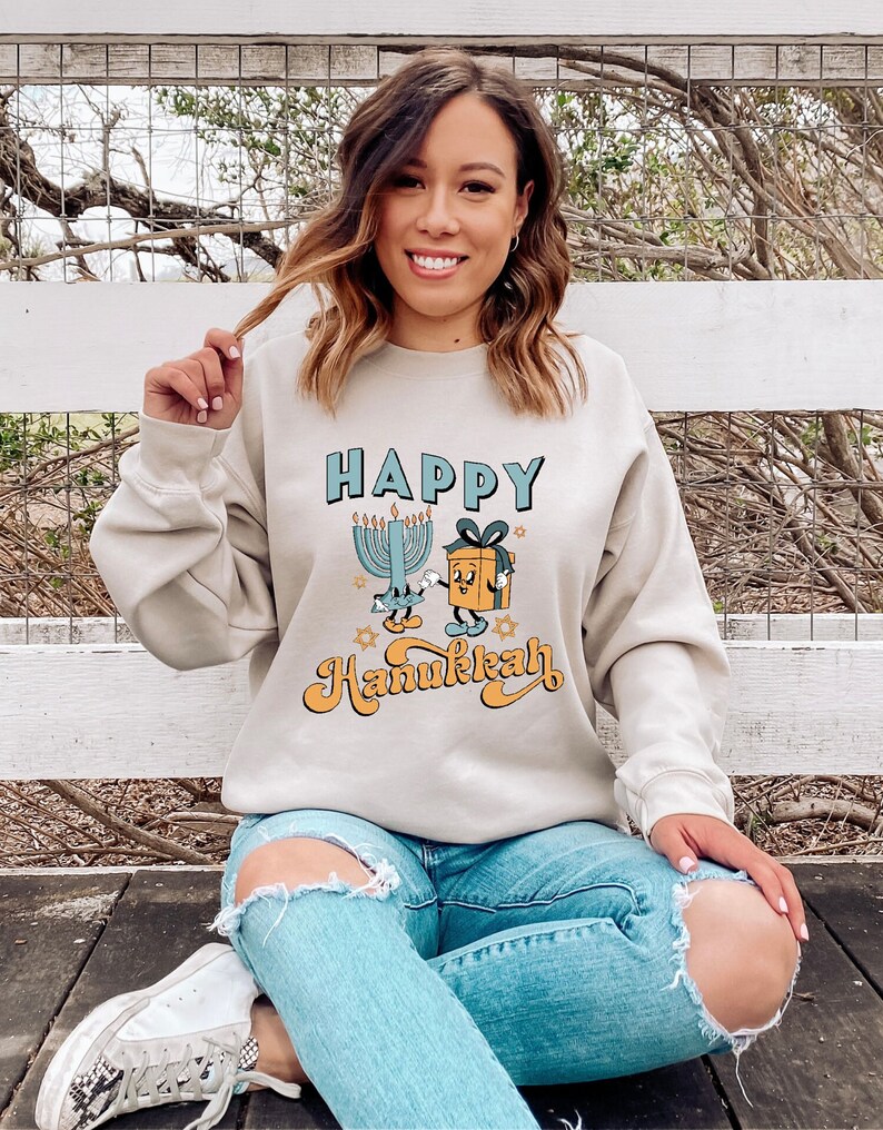 May include: Beige sweatshirt with the words "HAPPY Hanukkah" in blue and orange lettering. The graphic includes a cartoon menorah, a gift box, and stars. The person is wearing blue jeans and white sneakers.
