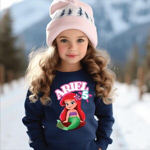 Może przedstawiać: Młoda dziewczyna w granatowej bluzie z grafiką Arielki z Małej Syrenki, z napisem "ARIEL" i cyfrą "5". Ma r&oacute;wnież na sobie jasnor&oacute;żową czapkę z białym wzorem drzew.