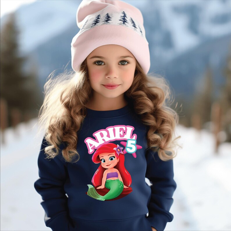 Może przedstawiać: Młoda dziewczyna w granatowej bluzie z grafiką Arielki z Małej Syrenki, z napisem "ARIEL" i cyfrą "5". Ma r&oacute;wnież na sobie jasnor&oacute;żową czapkę z białym wzorem drzew.