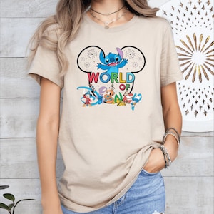 Pu&ograve; includere: T-shirt beige con una grafica colorata di Stitch di Lilo & Stitch, con la scritta "WORLD OF DISNEY". Il design include orecchie di Topolino con fuochi d'artificio e altri personaggi Disney.