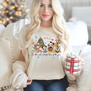 May include: Cream-colored t-shirt featuring a festive graphic of Disney characters, including Mickey Mouse, Donald Duck, and Pluto, surrounding a Christmas tree. The text "It's the most wonderful time of the year" is printed below the characters. The person is holding a red and white plaid mug.