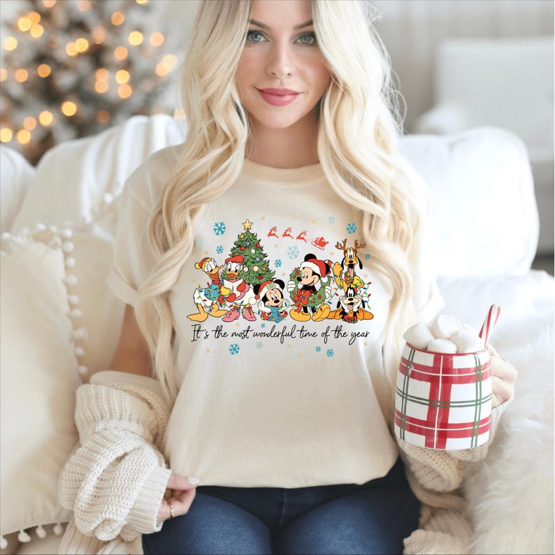 May include: Cream-colored t-shirt featuring a festive graphic of Disney characters, including Mickey Mouse, Donald Duck, and Pluto, surrounding a Christmas tree. The text "It's the most wonderful time of the year" is printed below the characters. The person is holding a red and white plaid mug.