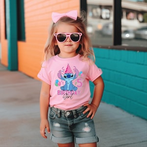 May include: A pink t-shirt with a blue cartoon character wearing a party hat and the number 5. The text "Birthday Girl" is also on the shirt. The child is wearing pink sunglasses and a pink bow.