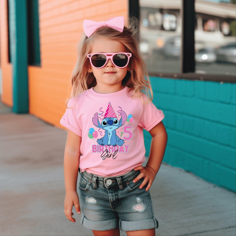 May include: A pink t-shirt with a blue cartoon character wearing a party hat and the number 5. The text "Birthday Girl" is also on the shirt. The child is wearing pink sunglasses and a pink bow.
