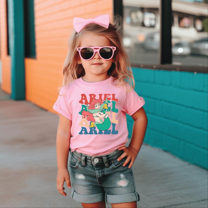 May include: A pink t-shirt featuring a retro-style graphic of Ariel the mermaid from The Little Mermaid. The graphic depicts Ariel with her signature red hair and green tail, surrounded by colourful text that reads "Ariel" multiple times.