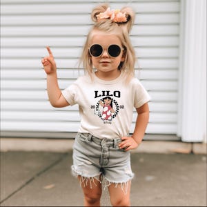 May include: A beige t-shirt featuring the word "LILO" in black, with a cartoon image of a character in a red dress. The shirt is worn by a young child wearing sunglasses and denim shorts.