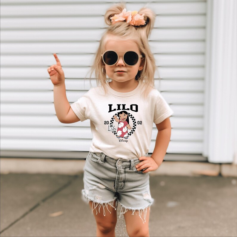May include: A beige t-shirt featuring the word "LILO" in black, with a cartoon image of a character in a red dress. The shirt is worn by a young child wearing sunglasses and denim shorts.