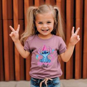 May include: A mauve t-shirt with a cartoon character wearing a party hat, balloons, and the text "5 Birthday Girl." The character is blue, and the text is pink and white. The shirt is worn by a young girl.