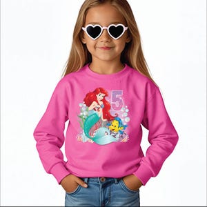 May include: Pink sweatshirt with a graphic of Ariel the Little Mermaid and Flounder. The graphic includes the number 5 and bubbles.